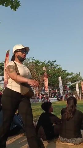 A video captures a sunny outdoor scene, possibly a park or festival, with people gathered. In the foreground, a woman with a tan complexion, wearing a white cap and sunglasses, is walking to the right, her hair tied back. She has a tattoo on her left arm and is wearing a beige t-shirt and black pants. A beige crossbody bag is slung over her right shoulder. To her left, a woman in a yellow shirt holds a young child wearing a denim dress and sandals. The child is looking forward with a curious expression. In the middle ground, a group of people are sitting on the grass, facing away from the camera, some appearing to watch an event. Tall, colorful banners are visible, adding to the festive atmosphere. A large tree with dense foliage stands behind the seated individuals, and beyond that, tents and more people suggest a lively gathering. The sky is clear and bright blue. In the right side of the frame, a young male in a black t-shirt and black shorts walks past, his body in profile.