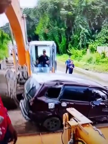 A maroon minivan is being lifted by an orange excavator arm. The excavator is positioned on a dirt road surrounded by lush green vegetation and trees. In the background, there are other vehicles and people standing on the road. The sky is overcast.