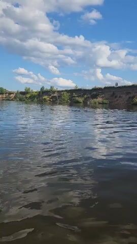 The video shows a wide body of water, likely a river, with a sandy bank on the opposite side. The bank is lined with green vegetation and trees. The sky is blue with fluffy white clouds. Several people are visible in the water, some appear to be swimming and others may be on inflatable devices. One person is standing on the bank at the far right of the frame. The camera appears to be moving slightly, possibly from a boat or while walking near the water's edge. The lighting suggests it is daytime and sunny.