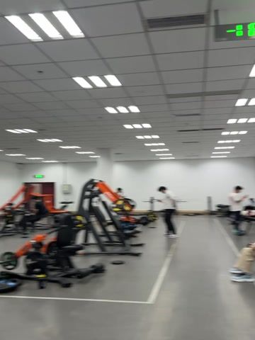 The video presents a gym environment with several individuals engaged in various activities. The gym features weight training equipment, including weight machines and barbells. Some individuals are actively working out, while others appear to be resting or using their phones. A man in a gray "BOSS" t-shirt is holding a 17.5 kg dumbbell. The gym is well-lit with overhead fluorescent lighting and has directional signage indicating exit routes.
