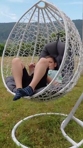 The video captures a young boy relaxing in a white, woven hanging chair outdoors. The chair is suspended by a chain from a stand and is situated on a grassy field with rolling hills and trees in the background. The sky is blue with scattered clouds, suggesting a pleasant, possibly warm day. The boy is wearing a light blue t-shirt, black shorts, and striped socks. He is seen interacting with his phone initially, then he looks up and smiles, appearing to enjoy himself. The camera pans around the chair, showing the boy's relaxed posture and the swinging motion of the chair. The setting appears to be a rural or semi-rural area in Chernyk, Ukraine, given the landscape. The overall mood is one of leisure and enjoyment. There is no visible text in the video.