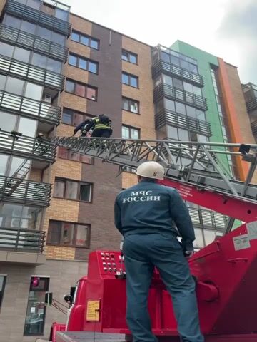 The video shows a rescue operation in front of a modern, multi-story apartment building. Two individuals, likely firefighters in uniform, are on an extendable ladder from a red fire truck. They appear to be assisting or rescuing someone from a balcony on an upper floor of the building. One rescuer is actively reaching out, while the other is positioned behind them. Another person in a similar uniform, wearing a white helmet, stands on the back of the fire truck, facing the building and observing the rescue. This person's uniform has text in Russian that reads "МЧС РОССИИ" which translates to "Ministry of Emergency Situations of Russia." The building has a mix of brick and colored facade elements, with balconies on most floors. The sky is overcast, suggesting it might be daytime but with subdued lighting. The scene depicts an emergency response with a focus on safety and rescue at height.