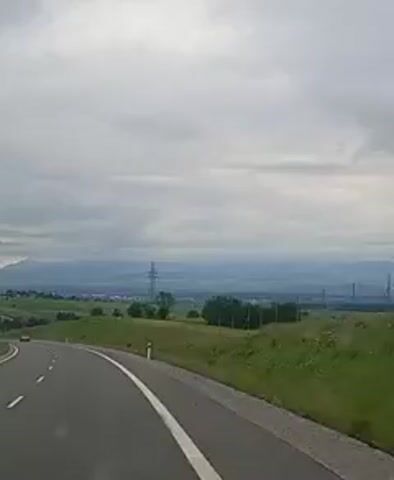 The video shows a highway with a curved road. The road is asphalt and has white lines marking the lanes. There are green grassy hills on the right side of the road, and a distant landscape with mountains and a power line tower on the left. The sky is cloudy and overcast. There are a few vehicles visible on the highway in the distance, including cars and a van. The perspective appears to be from inside a moving vehicle, likely a car.