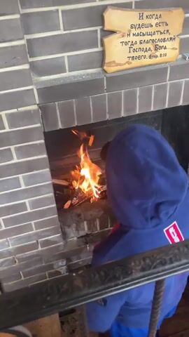 A child wearing a blue hooded jacket faces a fireplace. The fireplace contains burning logs and embers. Above the fireplace, a wooden plaque with text written in Cyrillic script is mounted on a brick wall. The child is holding a stick and appears to be interacting with the fire.