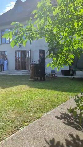 A person in dark clothing stands behind a wooden podium with some equipment on it in a grassy yard in front of a building. Another person in dark clothing walks past the podium. A young boy in dark clothing walks from the right side of the frame towards the left, pointing. Two people stand on a porch to the left of the building. The sun is shining, and leaves from a tree canopy cast shadows.