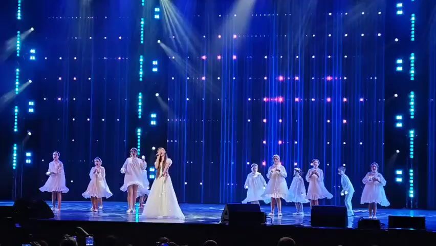 A young woman in a white gown stands center stage holding a microphone and singing. She is flanked by a group of children in white dresses who are dancing and holding candles. The stage is lit with blue and white lights, and the backdrop is a wall of vertical lights that change color. The audience is visible in the foreground, with many people holding up their phones to record the performance.