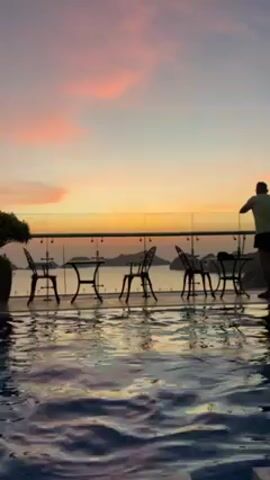 The video depicts an outdoor swimming pool at what appears to be a rooftop level during sunset. The sky is awash with colors of pink, orange, and yellow. A man in shorts and a light-colored shirt is standing near a glass railing, looking out at the view of the ocean and islands in the distance. Several tables and chairs are arranged near the pool. The water reflects the colors of the sky, creating a shimmering effect. The man eventually dives into the pool.