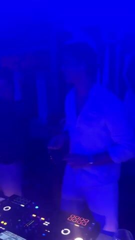 Two young men are in a dimly lit room with blue and purple lighting, likely a club or party venue, with DJ equipment visible in the foreground. The man on the right, wearing a white open-collared shirt and white shorts, is wearing sunglasses and appears to be dancing or gesturing enthusiastically with his hands. The man on the left, in a black shirt and white shorts, is also facing the DJ equipment and seems to be interacting with the equipment or the music. The background shows some decorative elements, including what looks like large cutouts or props.
