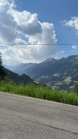 This is a vertical video showcasing a scenic mountain landscape in Ratschings - Racines, Italy. The video begins with a view of rolling green hills dotted with trees and some small buildings, with majestic, rugged mountains forming the backdrop. A paved road occupies the foreground, with lush green grass and wildflowers growing along its edge. A thin black wire stretches across the upper portion of the frame. The sky is a vibrant blue with fluffy white clouds, suggesting a clear, sunny day.

As the video progresses, the camera pans slightly, revealing more of the valley and the mountain range. The detailed textures of the asphalt road are visible, and the sunlight casts shadows, indicating it is likely daytime, possibly late morning or early afternoon. The overall impression is one of natural beauty and tranquility, evoking a sense of peace and awe. There is no visible text in the video. The scene suggests a drive through the mountains, possibly on a road trip, with the camera capturing the breathtaking views from inside or alongside a vehicle. The presence of a car's side mirror is visible at the very end of the clip, confirming the viewpoint of someone traveling by car.