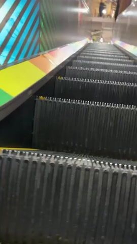 The video shows a first-person perspective looking down a black escalator with gray metal sides. The escalator steps are black with raised ridges. The sides of the escalator have a metallic finish with colorful striped designs featuring green, yellow, orange, and pink. At the bottom of the escalator, a store interior is visible with shelves of products and various signs, some with red and yellow accents. A person appears to be walking in the distance at the bottom of the escalator. The video is a continuous downward motion on the escalator.