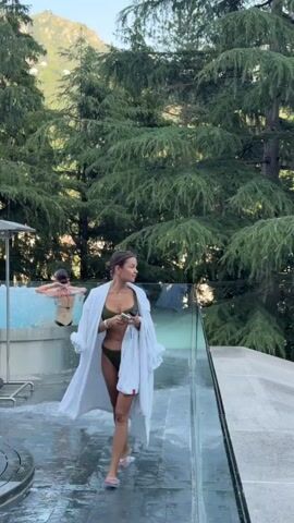 The video shows a woman in a green bikini and a white robe walking by an outdoor pool. She is holding a phone and a remote control. In the background, another woman is in the pool. The setting appears to be a resort with lush green trees and mountains in the distance. The camera pans out to reveal the entire pool area with lounge chairs and umbrellas.