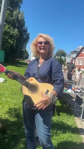 A woman with blonde curly hair and sunglasses stands holding a small guitar, smiling at the camera. She is wearing a dark blue top and jeans. In the background, there is a street with buildings, trees, and other people. The sun is shining brightly, indicating a clear day. The woman appears to be at an outdoor market or event.