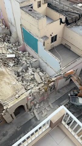 The video shows an excavator demolishing a building. The building is made of light-colored brick and has multiple levels. The excavator is orange and grey, and it is using its arm to break apart the building. There is a large pile of rubble on the ground in front of the building. The surrounding buildings are also made of brick and have a variety of colors, including white, pink, and blue. The video is shot from an elevated position, looking down on the demolition site. There is a white balcony railing in the foreground.