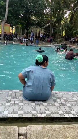 This media file shows a crowded outdoor swimming pool scene in Mlowokarangtalun, Indonesia. The pool is filled with people of various ages, most of whom are actively swimming or playing in the water. Several individuals are using inflatable rings, suggesting a recreational or leisure activity. In the foreground, a woman with her back to the camera is seated at the edge of the pool, wearing a blue shirt and a green hair accessory. Her posture suggests she might be resting or supervising.

The setting is characterized by lush greenery, with tall trees surrounding the pool, indicating a natural or semi-natural environment. In the background, there are structures that appear to be buildings or shelters, and more people are gathered, some sitting along the edge of the pool, possibly watching or waiting. The water is a vibrant turquoise blue, and sunlight is visible, suggesting it is daytime, likely during pleasant weather. The overall atmosphere appears lively and social, with many people enjoying the pool. There is no visible text in the media file.