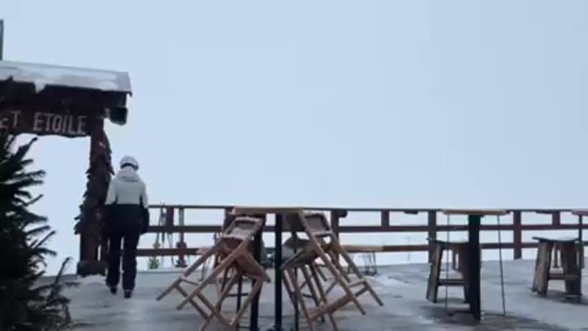 A person in ski gear is standing in front of a wooden sign that reads "CHALET ETOILE". It is snowing heavily. To the right of the person are several wooden tables and chairs, which are covered in snow. In the background, there is a railing and more snow-covered scenery. A pine tree is visible on the left. The person appears to be looking at their phone.