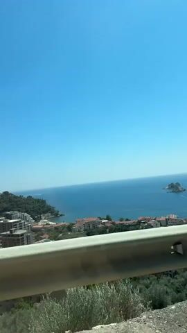 The video pans across a coastal landscape under a clear blue sky. In the foreground, a metallic guardrail is visible, with some greenery and rocky terrain below it. Beyond the guardrail, a town with red-tiled roofs and modern buildings is nestled on a hillside overlooking the sea. The blue ocean stretches to the horizon, with a small island or rocky outcrop visible in the distance. The camera is likely mounted on a moving vehicle, as evidenced by the slight motion blur and the changing perspective. The overall impression is a scenic view of a Mediterranean or similar coastal region.