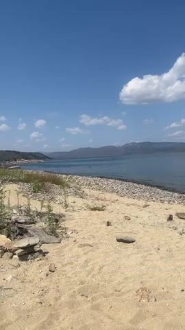 A still, eye-level, vertical video shows a sandy beach with scattered rocks and sparse vegetation. The beach leads to a calm, blue lake. In the distance, low, hazy mountains form the horizon under a bright blue sky dotted with white, puffy clouds. A red, rectangular object, possibly a floating device, is visible on the surface of the water in the middle distance. The scene is peaceful and appears to be a natural outdoor environment.