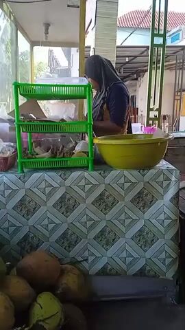The video shows a woman in a hijab standing at a food stall. She is wearing a dark blue shirt and yellow apron. In front of her is a table covered with a patterned tablecloth. On the table is a green plastic three-tier shelf with various items, including what appears to be fried food, and a large yellow bowl. Beneath the table are several coconuts. The stall is partially enclosed with a glass front. Outside, there is a building with a red tile roof and a green metal structure. The woman appears to be preparing or organizing food at the stall.