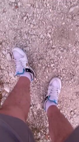 The video begins with a close-up shot of a person's legs and feet wearing white athletic shoes and grey shorts, standing on a gravelly surface. The camera then pans to the right, showing a landscape of rolling hills and mountains under a twilight sky with pink and purple hues. A woman in black athletic wear, holding a smartphone, is visible in the mid-ground, taking a selfie. The video concludes with sweeping views of the mountainous terrain and the colorful sky.