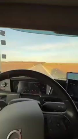 The video shows a first-person view from inside the cabin of a truck driving on a straight, rural road on a sunny day. The road stretches into the distance between fields that appear to be harvested or fallow.  The truck's dashboard is visible, including the steering wheel, instrument panel showing a speed of approximately 90, and a navigation/entertainment display showing music is playing. A GPS device is also mounted on the dashboard. The sky is clear with a few scattered clouds. The overall scene is peaceful and suggests a long-distance drive.