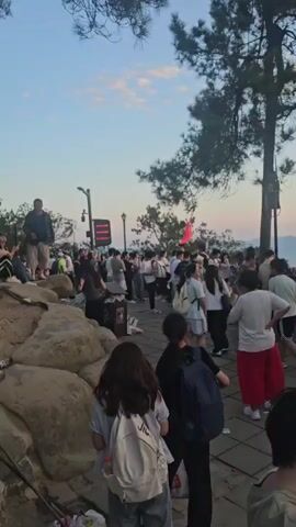 The video shows a crowd of people gathered outdoors on a paved area with a stone wall. Several large trees with pine needles are present. In the background, a hazy mountainous landscape is visible under a pale sky, suggesting sunset or sunrise. Some people are standing and looking in various directions, while others appear to be engaged in conversation. A red flag is visible in the distance, held by someone in the crowd. There are some trash cans and scattered debris on the ground. The overall atmosphere seems casual, with people dressed in everyday clothing.