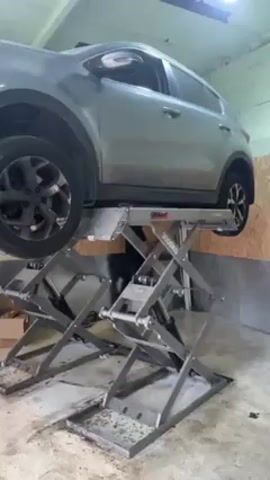 The video shows a dirty grey Kia Sportage raised on a grey hydraulic scissor lift. The lift is positioned in a garage with painted concrete walls and floor. The garage also contains boxes and wood paneling. The lift is in operation and is holding the Kia Sportage off the ground.