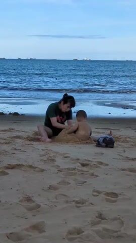This media file shows two people on a beach. A woman wearing a dark green t-shirt is sitting on the sand, facing a young child who is partially buried in a mound of sand. The woman is using her hands to shape the sand, and the child is looking towards her. The child is wearing a light-colored diaper or shorts. To the right of the child, a dark-colored cloth or bag is lying on the sand.

The scene is set on a beach with a shoreline and the ocean in the background. The water is a deep blue, and there are several ships visible on the horizon. The sky is a light blue with some clouds. The sand on the beach is light brown and shows imprints of footsteps.

The overall impression is one of a casual, family-friendly outing at the beach. The time of day appears to be late afternoon or early evening, given the soft lighting and the slight haziness in the sky. The weather seems pleasant, suitable for beach activities.

The t-shirt worn by the woman has some text on it, which appears to be green and possibly in a stylized font, but it is difficult to read clearly. The activity depicted is playing in the sand, a common pastime at the beach, suggesting a relaxed and enjoyable atmosphere. The emotions conveyed are likely happiness and engagement, with the woman interacting with the child. The location cues, such as the ocean and the presence of ships on the horizon, are consistent with a coastal setting, and the provided context places this scene in Quy Nhon, Vietnam.