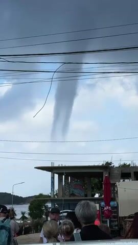 The video captures a group of people gathered in what appears to be a coastal town, looking up at a distinctive funnel cloud forming in the sky. The scene is shot from behind the spectators, with power lines crossing the top of the frame.

In the foreground, several individuals, mostly visible from the shoulders up, face away from the camera, their attention directed upwards. Some appear to be holding phones, possibly recording the event. Below the power lines, the roofline and upper structure of a concrete building are visible, showing exposed rebar and graffiti on its walls, suggesting it might be under construction or disused. A red umbrella and a cooler or display stand with partial text "RRTA" are also seen to the right of this building.

The mid-ground features the tops of palm trees and other greenery. In the distance, a body of water, likely the sea, is visible, confirming the coastal setting (consistent with Ksamil, Albania).

The dominant feature is the funnel cloud, a prominent, elongated vortex of clouds descending from the sky. It appears as a dark, column-like formation against a brighter, partly cloudy sky. A thin, dark, curved line, possibly a wire or string from a kite, is also visible near the funnel cloud. The lighting suggests it is daytime, under overcast conditions. The overall atmosphere is one of observation and perhaps wonder or concern as people witness the unusual weather phenomenon.