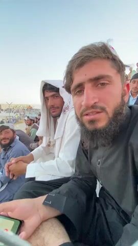 In the foreground, a man with a beard and wearing a black tunic is holding a smartphone and looking at the camera. To his left, another man with a white head covering is looking forward. To the left of him, a third man in a blue outfit is looking to his left. In the background, a crowd of people can be seen sitting and standing in what appears to be an outdoor venue. The sky is clear and bright.
