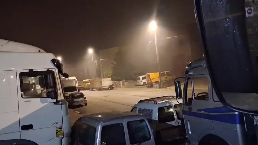The video captures a nighttime scene in an outdoor area, likely a parking lot or industrial zone, with several vehicles present. The lighting is dim, primarily from streetlights and possibly interior lights of the vehicles. A large white truck dominates the left side of the frame, with its cabin visible. To its right, a smaller silver car is parked. Further into the scene, there are more trucks and vans, including a yellow van and a white box truck. The ground appears to be paved. There is a noticeable haze or fog in the air, illuminated by the streetlights. The right side of the frame shows the reflection in a mirror, possibly from another vehicle, obscuring some of the scene. The overall atmosphere is industrial and somewhat desolate due to the time of day and limited lighting. The audio includes muffled speech and what sounds like the engine of a vehicle.