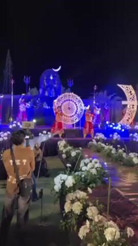 At night, a group of four young dancers, adorned in sparkling red costumes, perform on a stage brightly illuminated with blue lights. In the background, a large, shimmering statue of a head is placed under a crescent moon, while decorative floral arrangements line the stage's edge. A videographer records the performance from the side, and the scene suggests a celebration of faith or cultural identity.