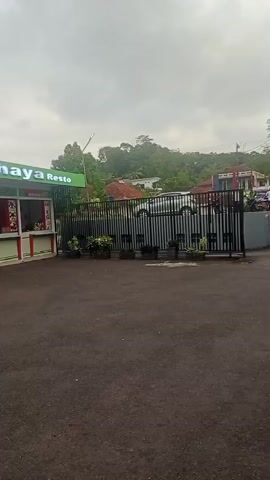 The video shows an outdoor food court area. A sign above the food stalls reads "Foodcourt Cahaya Resto". There are several food stalls with customers being served. To the right of the food stalls, there is a black metal fence with potted plants in front of it. Beyond the fence, several cars are parked. A group of people are standing and walking near the parked cars. The sky is cloudy. The ground is paved.
