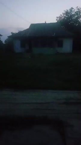 The video is a low-light recording of a house situated on a grassy incline. The house is single-story with a dark, sloped roof. A porch runs along the front of the house. Trees are visible behind and to the right of the house. The foreground of the video shows a dark, textured surface, possibly a road or path. The sky above is a pale blue, suggesting dusk or dawn. No people or explicit actions are clearly discernible due to the low light conditions.