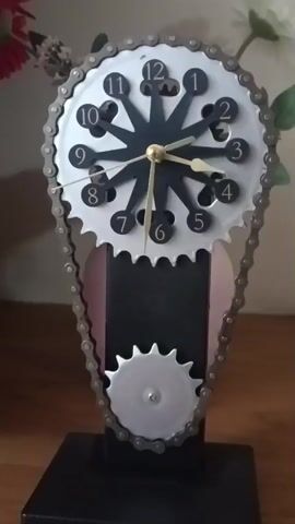 A close-up video captures a unique tabletop clock designed with bicycle gear and chain elements, set indoors on a wooden surface. The clock features a large silver gear as its face, encircled by a metal chain that extends down to a smaller silver gear mounted on a black stand. The clock face has a black star-shaped center with silver numbers 1 through 12, each within a separate circular cutout on the gear. Gold-colored hour and minute hands, along with a yellow second hand, indicate the time. The second hand is visibly sweeping. The time displayed is approximately 1:53. The background is softly blurred, showing glimpses of artificial foliage, including green leaves and reddish flowers. The clock itself rests on a sturdy black base.