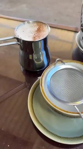 The video opens with a close-up of a metal pot with a handle containing a frothy beverage, possibly coffee or tea, dusted with cinnamon. The pot sits on a dark, reflective surface, likely a table. A small metal strainer is placed on top of a stack of saucers and a cup. The camera then pulls back slightly to reveal more of the scene. To the right of the cup and strainer, there is a potted plant with green leaves and a decorative pot. The background appears to be outdoors, with natural light suggesting daytime. The video then continues to pan and zoom slightly, offering different perspectives of the same setup, emphasizing the beverage, the strainer, and the plant. There are no people visible in the video. The overall setting suggests a moment of preparation or enjoyment of a hot drink.