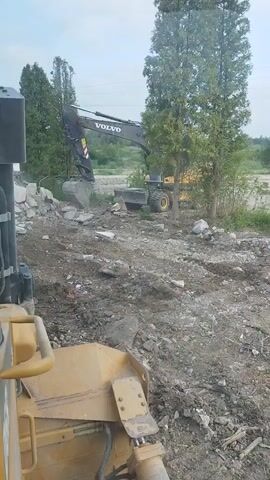 The video shows a construction site with two excavators. The foreground features a yellow bulldozer. In the background, a black Volvo excavator is actively demolishing a pile of concrete debris. The excavator's arm is extended, and its bucket is scooping up chunks of concrete, causing dust to rise. Another yellow piece of heavy machinery, possibly a wheel loader, is parked behind the Volvo excavator. The ground is covered in dirt, rubble, and some green vegetation. Tall trees are visible in the background, and the sky appears overcast. The overall scene depicts active demolition and construction work.