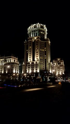 The image displays a large, ornate building at night, illuminated by numerous warm lights. The building is grand in scale, featuring classical architectural elements such as prominent columns, multiple stories, and a distinctive rounded or dome-like structure at its pinnacle, which is also brightly lit. Numerous windows across the facade show internal lights, suggesting occupation.

The sky is completely dark, emphasizing the brightness of the building's exterior lighting. To the right of the main building, a street or open area is visible, with some ground lighting casting a glow. Other, less prominent illuminated buildings or structures can be seen in the background, extending into the distance. The overall impression is one of a prominent landmark or hotel beautifully lit up in an urban setting after dark.

The image itself is oriented vertically, as if the camera was held on its side, but the content is clearly a horizontal scene. When viewed with the building upright, it stands tall against the dark night sky.