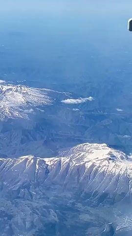 This aerial view captures a vast and rugged mountainous landscape under a bright, clear daytime sky. The scene is dominated by multiple mountain ranges, with several prominent peaks covered in glistening white snow, indicating either high altitude or a cooler season. The snow highlights the sharp, irregular contours of the mountain ridges.

Deep valleys and lower-lying hills, covered in shades of green and brown, separate these majestic formations. Scattered small, white clouds are visible, both above the peaks and hovering within some of the valleys. In the distance, the landscape fades into a soft blue haze, characteristic of atmospheric perspective from a high altitude.

Evidence of human activity is subtly present, with winding roads and small clusters of buildings, likely villages or towns, nestled within the valleys and at the foothills of the mountains. A portion of an aircraft's wing is visible in the top right corner, indicating the high-altitude perspective from a plane. The overall impression is one of natural grandeur and tranquility. The location context provided is Rocca di Mezzo, Italy, which aligns with the visual characteristics of the Apennine Mountains. There is no visible text in the image.