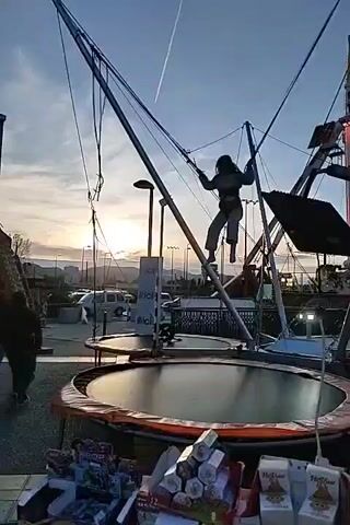 The video shows a carnival with several attractions. A woman is jumping on a bungee trampoline, attached to wires and performing flips. The camera pans to a large pendulum ride, which is decorated with lights and is swinging back and forth. There are people standing in line to ride the pendulum. The scene is set against a backdrop of buildings and a sunset.