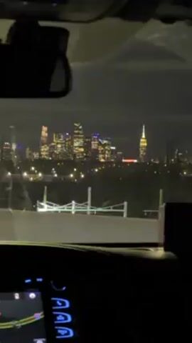 The video shows a view of the New York City skyline at night, as seen from inside a moving vehicle. The Empire State Building is visible, along with other illuminated skyscrapers. The car's dashboard and rearview mirror are also visible, suggesting the video was taken from the driver's or passenger's perspective. The environment appears to be a highway or elevated road, with guardrails in the foreground.