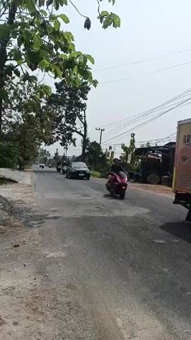 A red motorcycle with a black seat and a license plate is seen from behind on an asphalt road. The motorcycle is ridden by a person wearing a black helmet and black clothing. In the background, several other motorcycles are visible, some with riders, on the same road. A dark-colored car is also on the road. Trees with green leaves line the left side of the road. The sky is clear and bright, suggesting daytime. The road surface appears somewhat worn and uneven. There are utility poles and wires visible in the background, along with some buildings and vegetation. The scene appears to be in a rural or suburban area.