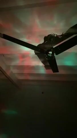 A ceiling fan is visible in the center of the frame, its blades rotating. The ceiling and walls are illuminated by colorful lights that project patterns resembling nebulae and stars. The colors shift and change, from red and green to green, and then to blue. The room is dark, with the fan and the projected lights being the primary sources of illumination. The camera is pointed upwards, capturing the fan and the light display on the ceiling.