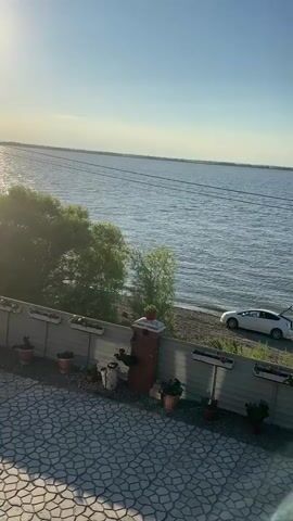 A white car with its trunk open is parked on a gravelly shoreline next to a body of water. A person is sitting on a chair next to the car, facing the water, with fishing rods nearby. In the foreground, a paved area with a decorative pattern is visible, along with a fence lined with potted plants. The sky is clear and blue.