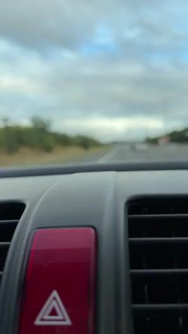 The media file captures a first-person perspective from inside a car, looking forward through the windshield. The primary subjects are the interior of the car and the road ahead. In the foreground, a red hazard light button with a white triangle symbol is prominently visible, situated above a car's air vent.

The scene outside the car depicts a road winding through a landscape. Green trees and foliage line the sides of the road, suggesting a rural or semi-rural setting. The road itself appears to be a highway or a main road, with other vehicles visible in the distance, creating a sense of ongoing travel. The sky is overcast with dramatic clouds, indicating a day with changeable weather, possibly late afternoon given the lighting.

The activity taking place is driving. The focus on the hazard light button might imply a need for caution or a specific driving situation. The overall mood conveyed is one of travel and perhaps a touch of introspection due to the solitary perspective and the somewhat dramatic sky.

There is no visible text within the media file itself, apart from the symbol on the hazard light. The location context provided places this scene in the Malvern Hills, United Kingdom, which aligns with the lush green scenery. The time of day is not precisely determinable but appears to be daytime.