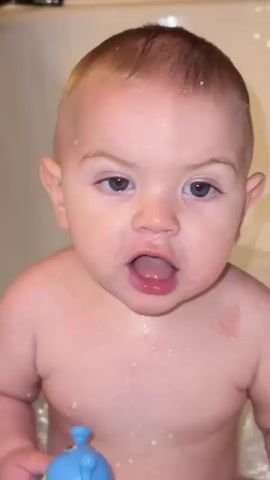 A toddler with light hair and blue eyes, wearing no clothes, stands in a bathtub. They are covered in water droplets. The toddler grins and then laughs, biting a blue toy. The bathtub is white with dark brown tiles above it.