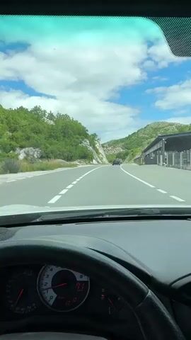 A first-person perspective video shows a car driving on a highway through a mountainous, green landscape. The driver's side dashboard is visible, including the steering wheel, speedometer, and tachometer. The speedometer reads 71-72. The road ahead curves slightly, with white lines marking the lanes. On either side of the road are steep, rocky hillsides covered in lush green trees and vegetation. The sky is blue with scattered white clouds. In the distance, another vehicle is visible on the road. To the right of the road, there are what appear to be structures with a patterned roof and signs. The overall impression is of a scenic drive in a natural setting.