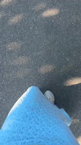 The video shows a close-up, top-down view of a person's feet and lower legs as they walk on asphalt. The person is wearing white sneakers and a long, light blue dress with a textured, almost lace-like pattern. Sunlight casts dappled shadows on the asphalt in front of the person. The camera angle suggests the video is being recorded by the person walking, likely with a phone held out in front of them. The movement is smooth, indicating a steady walking pace. The environment appears to be outdoors on a paved surface, possibly a sidewalk or street.