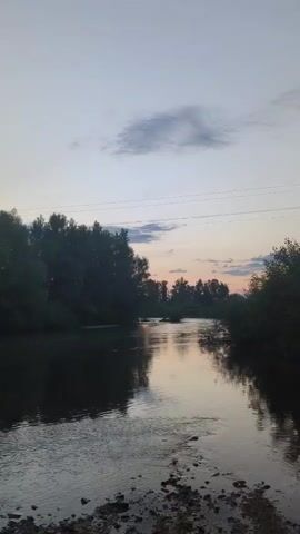 The video depicts a tranquil scene of a river at dusk, with trees lining its banks. The camera pans across the water, capturing the reflection of the sky and trees. In the background, power lines are visible, stretching across the landscape. The sky is filled with clouds, and the soft hues of the setting sun are reflected in the water, creating a peaceful and serene atmosphere. The video has no audio.