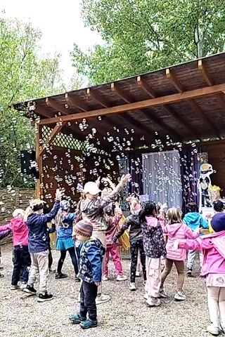 A group of young children are gathered in front of a wooden stage or pavilion, reaching up towards a multitude of soap bubbles floating in the air. The children are of various ethnicities and wear diverse clothing including jackets, hats, and brightly colored outfits. The stage has a dark wooden roof, lattice details, and a silver curtain backdrop. Two figures stand on the stage, one slightly obscured. The ground beneath the children is covered with gravel. Green trees are visible in the background, indicating an outdoor setting. The overall atmosphere is joyful and playful, accompanied by music.