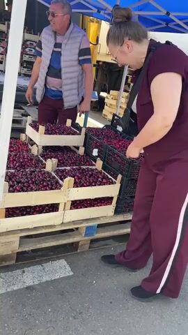 A man wearing sunglasses and a vest is standing behind a stall filled with crates of cherries. A woman in a burgundy tracksuit is standing in front of the stall, looking at the cherries. She is wearing glasses and has a black fanny pack. In the background, a yellow truck is parked with its back door open, revealing more crates of produce. Another man is sitting on a pallet of boxes near the truck. The scene appears to be an outdoor market.